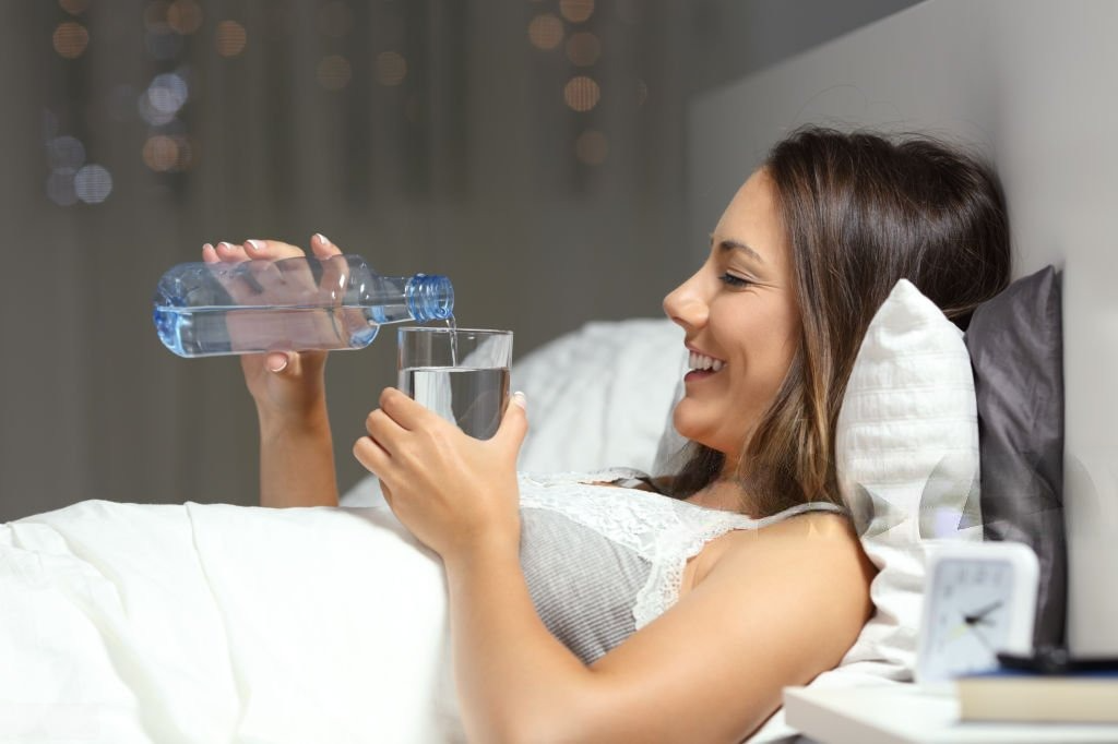 a woman lying in bed pouring water into a glass. Drinking Water Before Bed