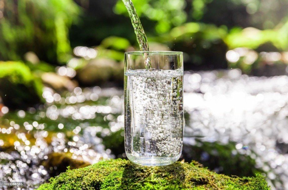  Drinking Water RO System, A close-up of clean purified water filling in a drinking glass, placed on a moss-covered rock with a flowing river in the background in a forest setting.