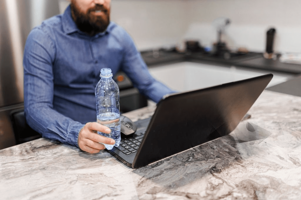 A man drinking water from a glass while working on a laptop in the kitchen, illustrating the importance of hydration during remote work.